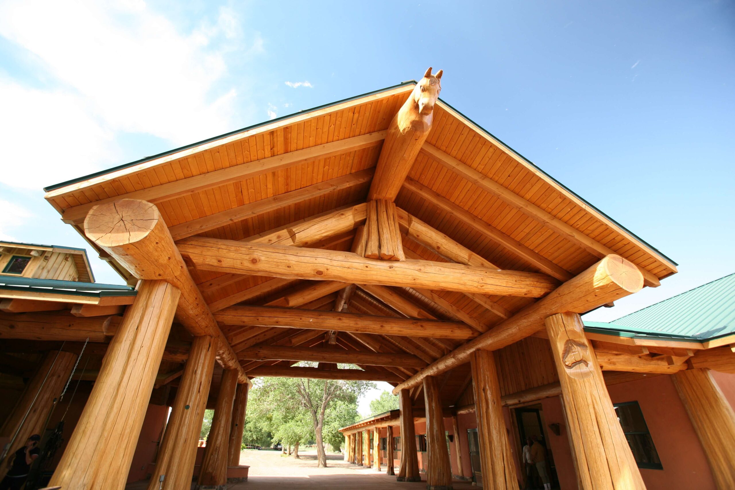 Hauptportal eines Reitstalls mit massiven Holzsäulen und einem Pferdekopf-Schnitzerei, der über den Eingangsbereich blickt, unterstrichen durch die offene Architektur und das natürliche Holzdesign